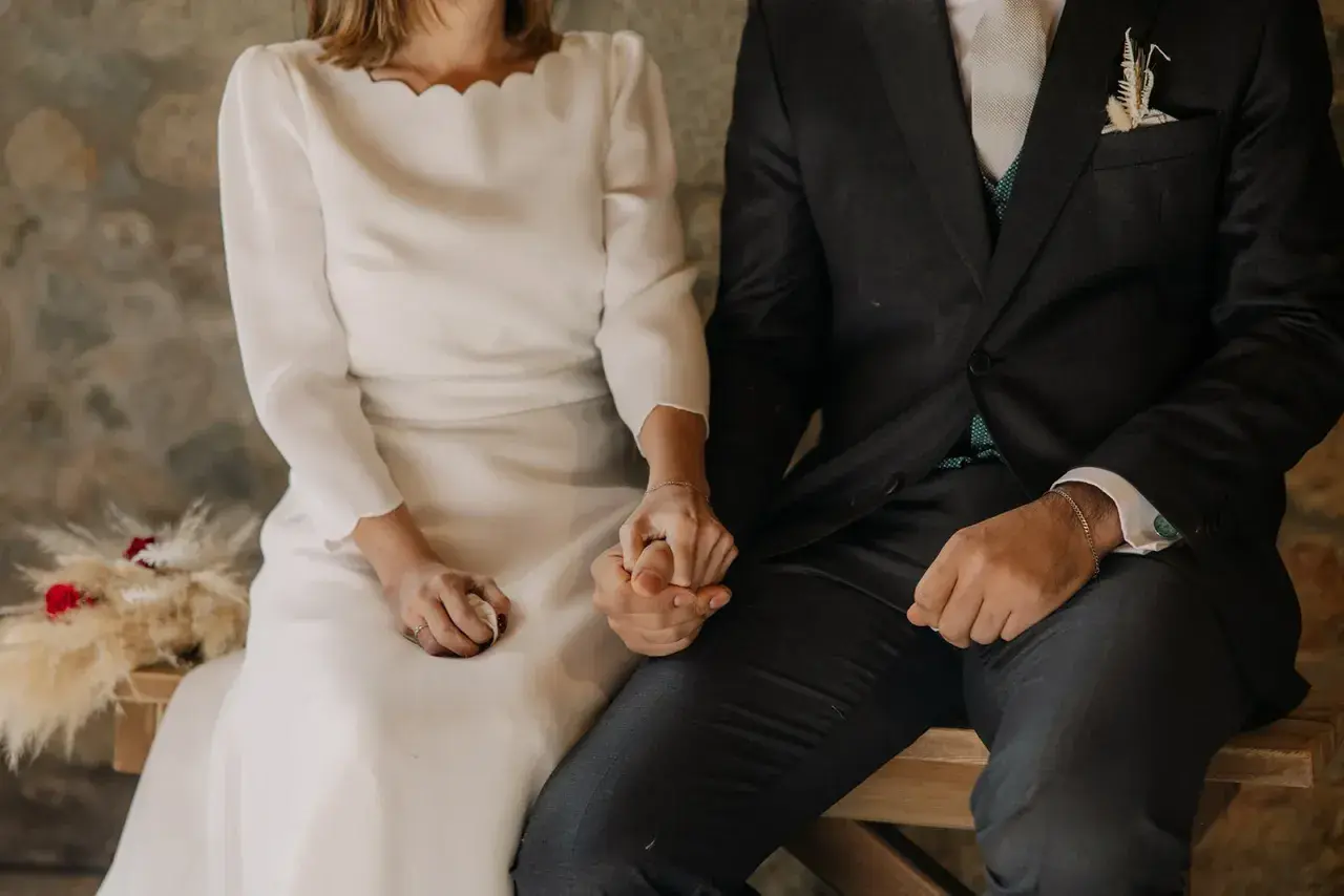 Novios tomados de la mano, ella con vestido blanco y él con traje oscuro. Se ve un anillo casado en su mano.