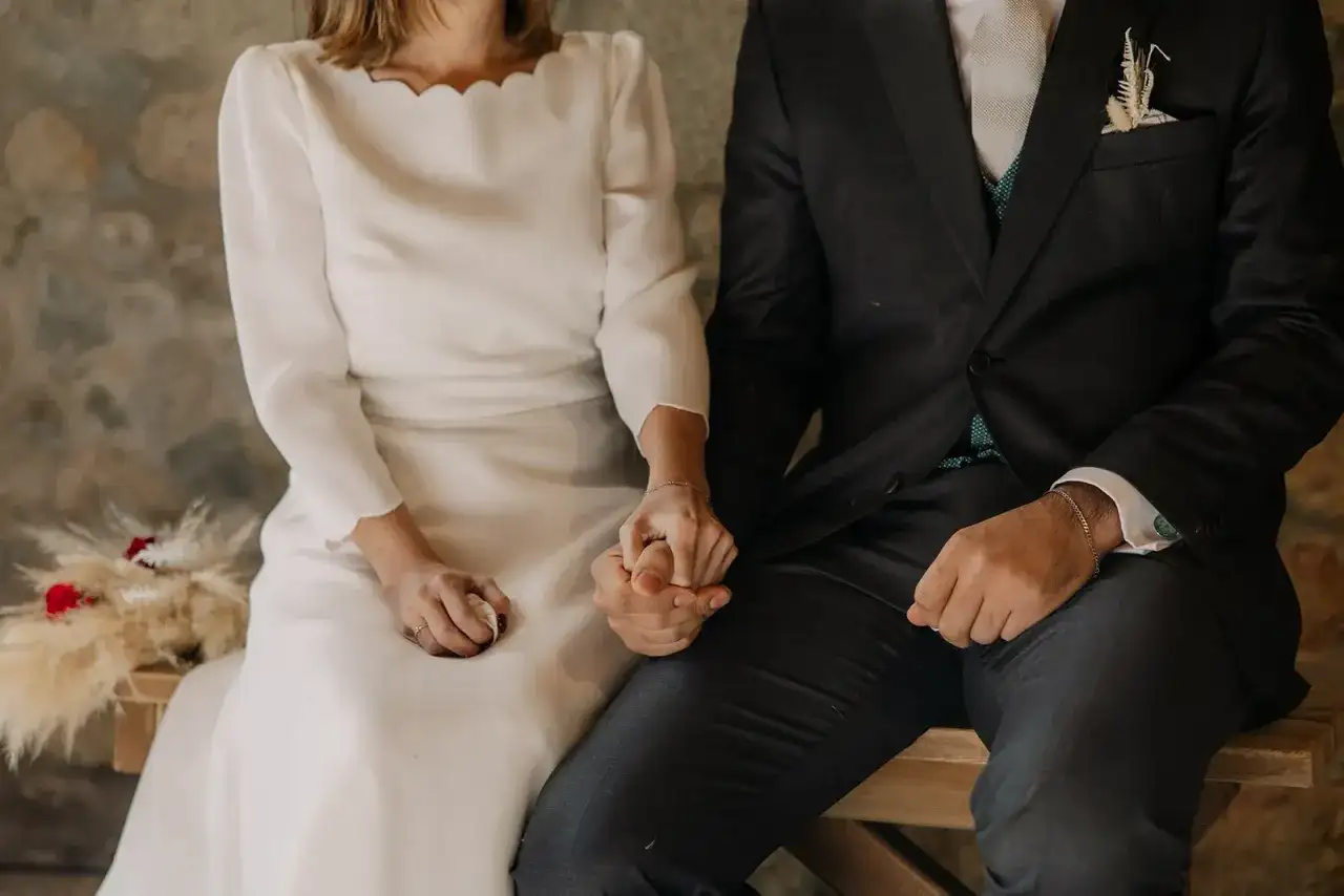 Novios tomados de la mano, ella con vestido blanco y él con traje oscuro. Se ve un anillo casado en su mano.
